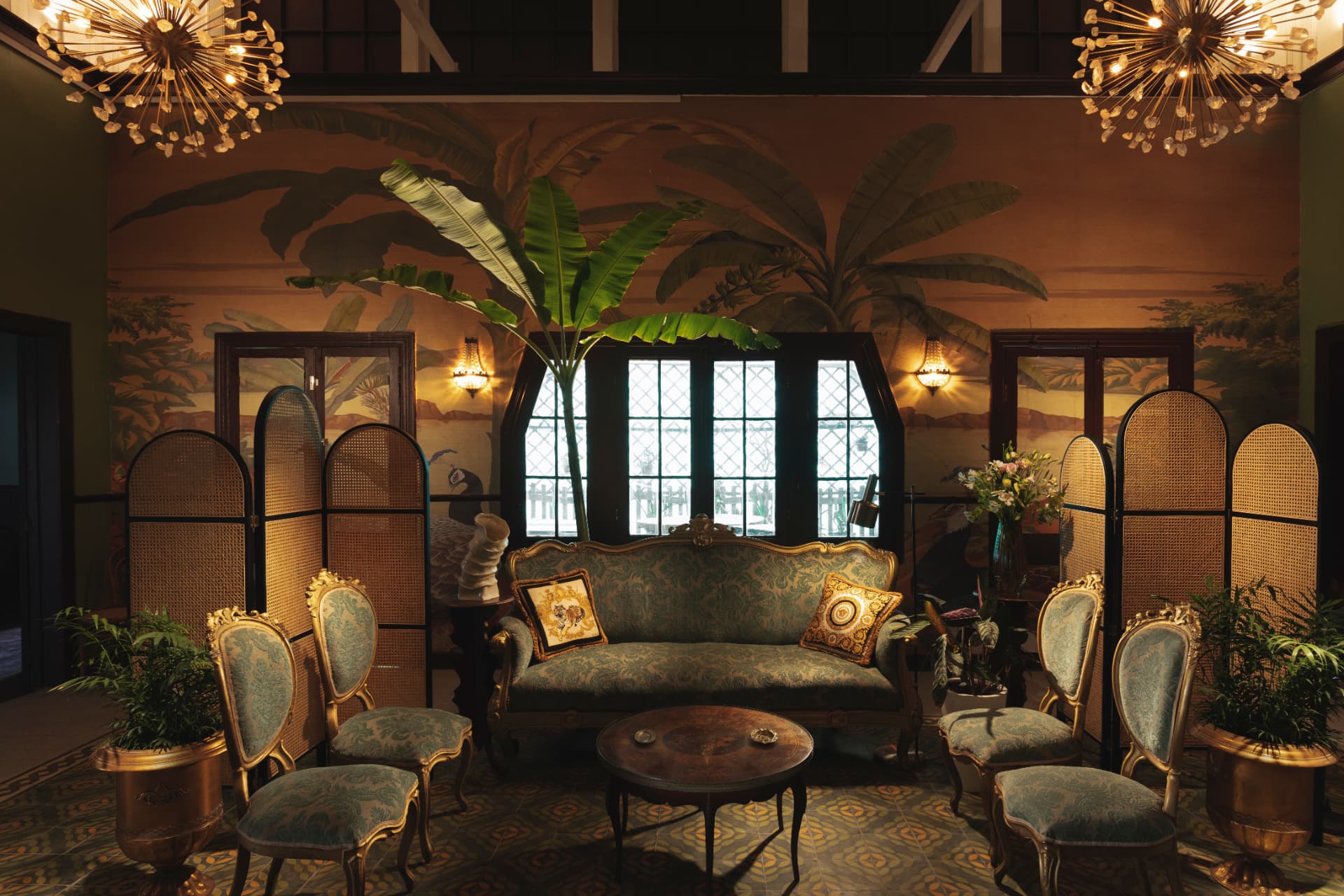 Gran salón de Casa Vallereggia con sofá antiguo, mural tropical y lámparas encendidas